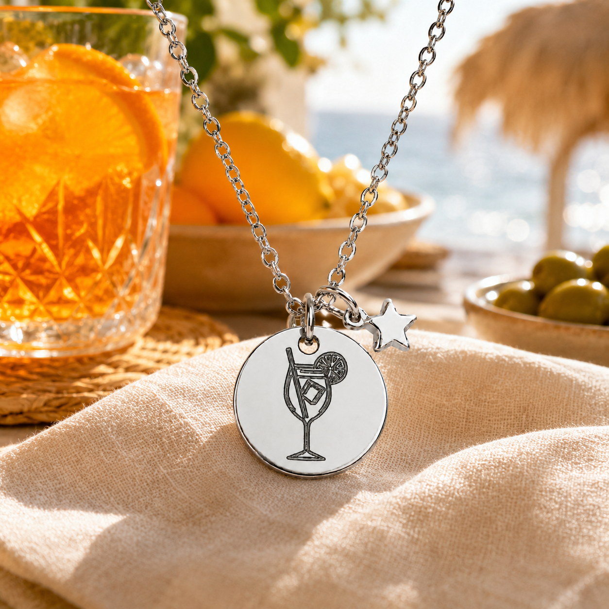 Sommerliche Edelstahl-Halskette mit Cocktail-Anhänger und Stern-Charm auf hellem Stoff am Strand mit Orangen und Sommerdrink im Hintergrund.