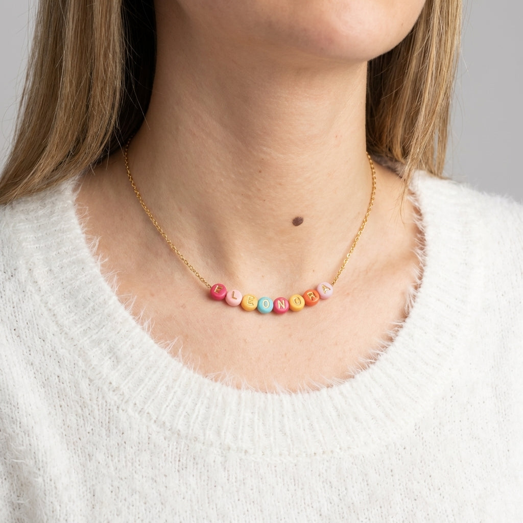 Frau trägt eine goldene Namenskette mit bunten Buchstaben-Perlen (Eleonora) auf weißem Pullover vor neutralem Hintergrund